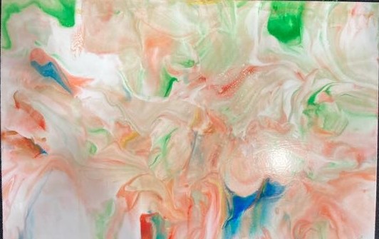 A close-up shot of a piece of photo paper with swirling, abstract colors. There are reds, blues, greens, and yellows, but the most prominent color is the reddish-pink color that takes up most of the paper. It rests on a black fabric background.